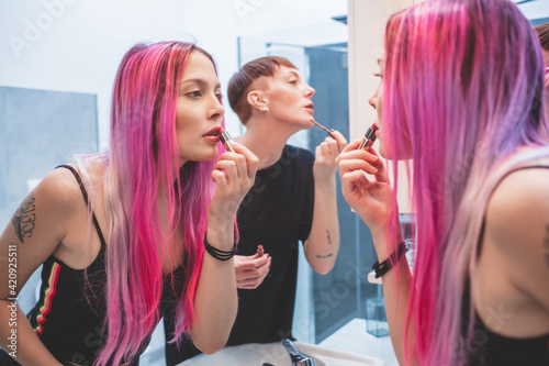 Young woman with long pink hair and woman with short red hair standing in front of mirror, applying lipstick.
