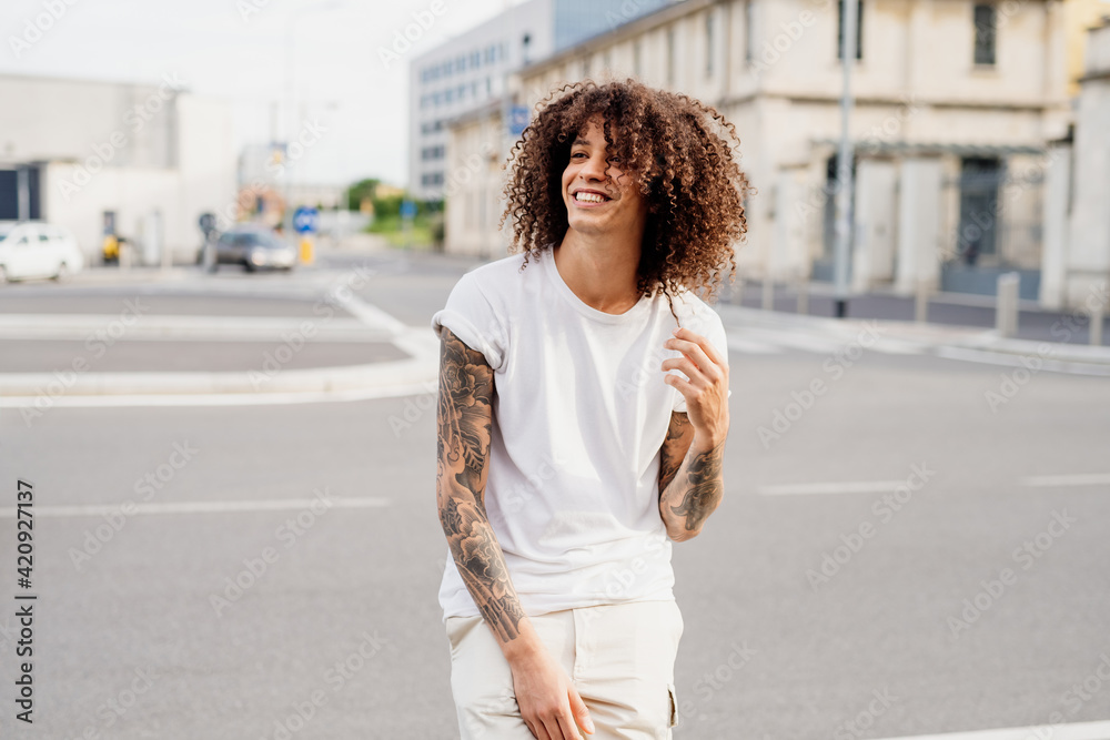 Smiling man with tattooed arms and long brown curly hair standing on a street.