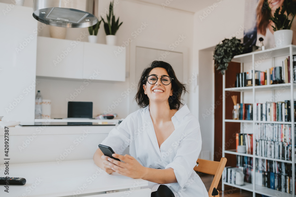 Woman with brown hair wearing glasses sitting at table in an apartment, smiling at camera. Woman with brown hair wearing glasses sitting at table in an apartment, smiling at camera.
