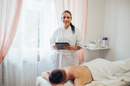 Smiling beautician standing next to woman lying on treatment bed in beauty salon.