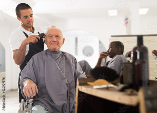 Elderly man getting haircut with electric clipper from skillful young barber in salon..