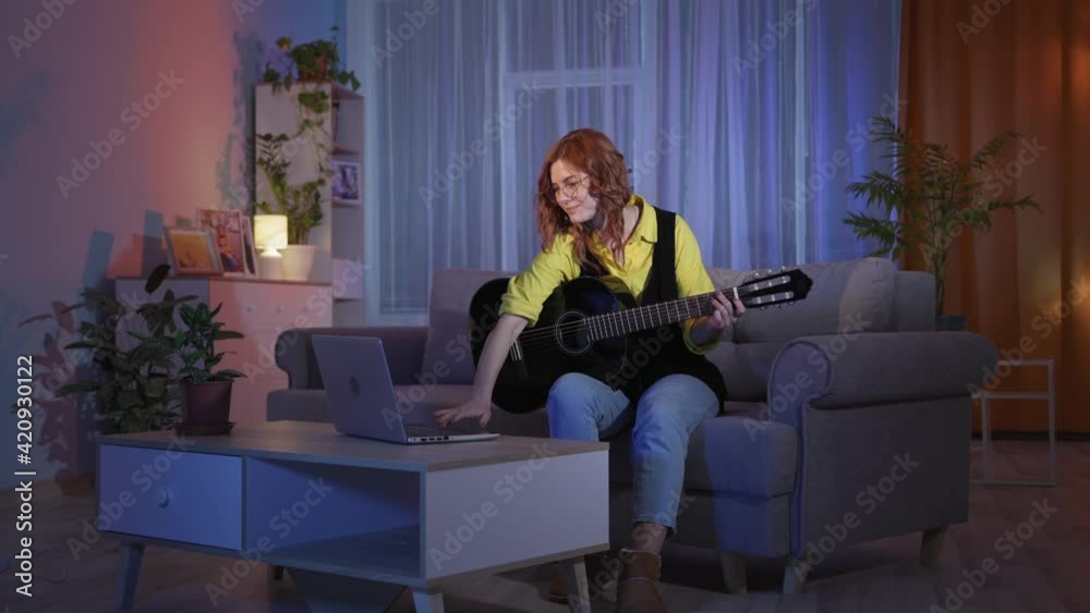 young woman with glasses playing acoustic guitar learning notes remotely using laptop while sitting on couch in evening at home young woman with glasses playing acoustic guitar learning notes remotely using laptop while sitting on couch in evening at home