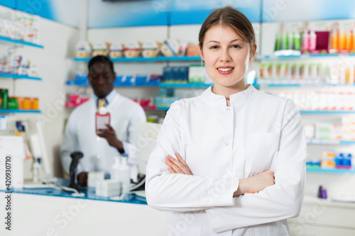 Experienced cheerful positive smiling female pharmacist standing on background with male colleague in pharmacy