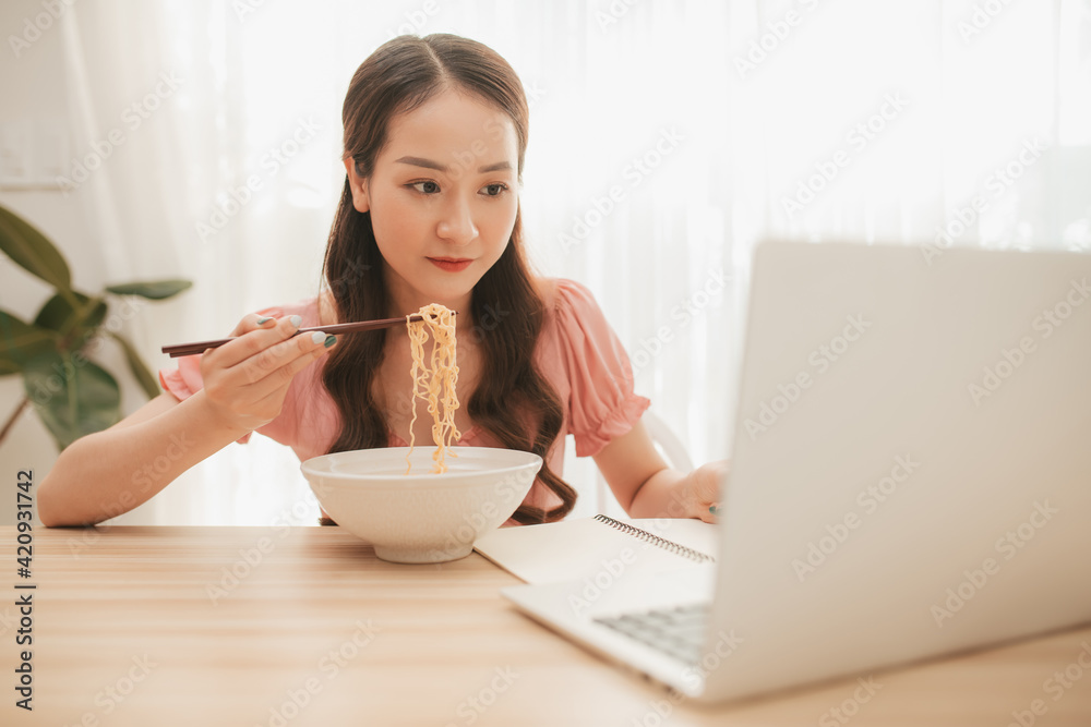 Girl eating instant noodle in hurry time while running her business at home on notebook computer Girl eating instant noodle in hurry time while running her business at home on notebook computer