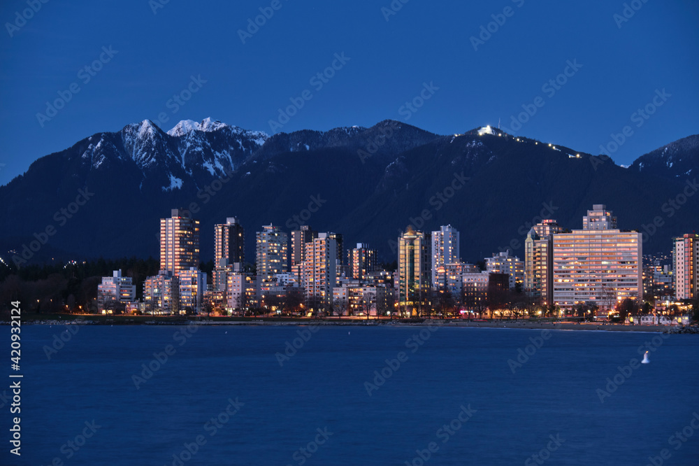 Fototapeta premium Highrise buildings by ocean and mountains at dusk. West side Vancouver and grouse mountain from Kitcilano Beach. British Columbia. Canada