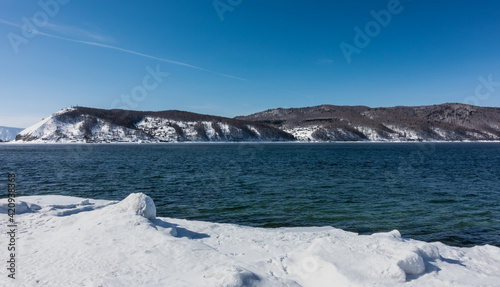 Never freezing source of the Angara River. Blue water flows between the snow-covered shores. In the distance  against the background of the sky  mountains covered with snow. Russia