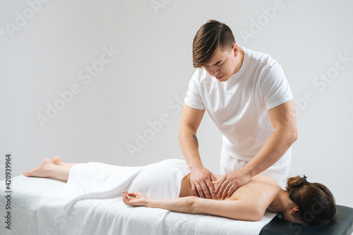 Side view of male masseur massaging lower back of young woman lying on massage table on white background. Professional physiotherapist performing wellness treatments for female with back pain.