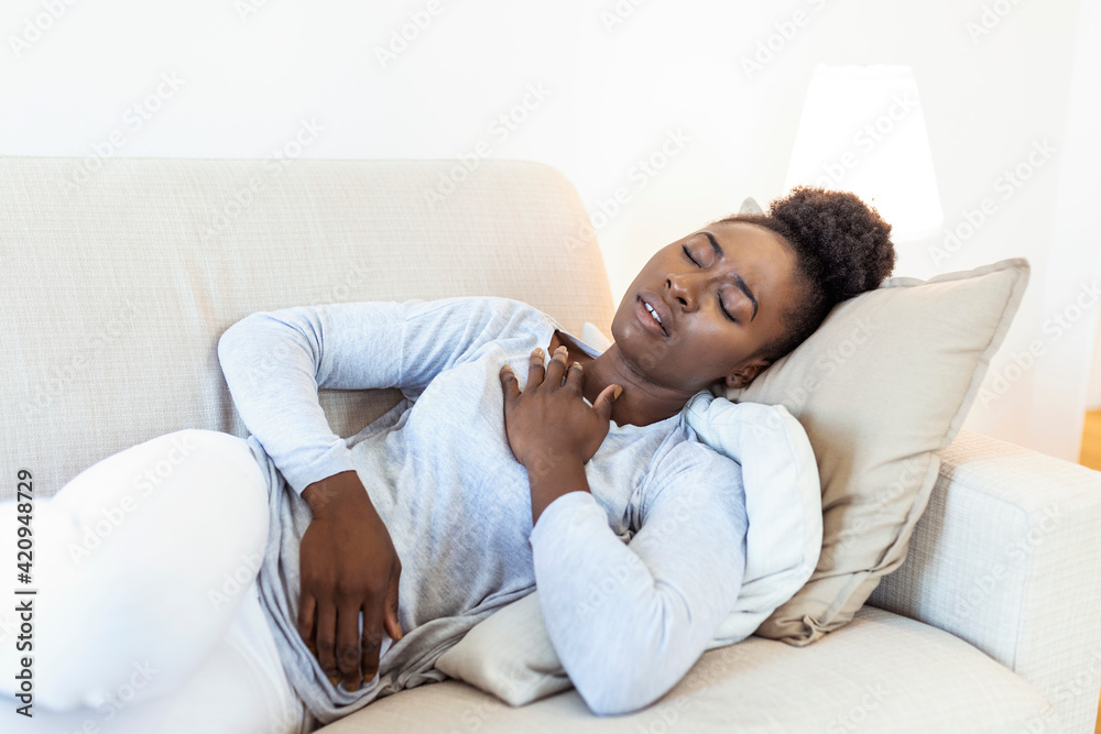 African beautiful woman sick with feeling pain in the chest while laying on the sofa .Hand holding chest after taking a medication.Concept of hard work without maintaining health. African beautiful woman sick with feeling pain in the chest while laying on the sofa .Hand holding chest after taking a medication.Concept of hard work without maintaining health.