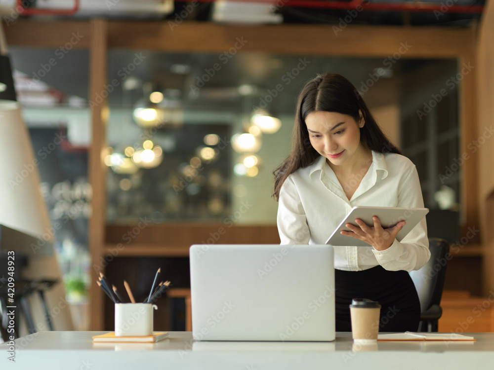 Female office worker working with laptop and digital tablet on her hand while standing at office desk Female office worker working with laptop and digital tablet on her hand while standing at office desk