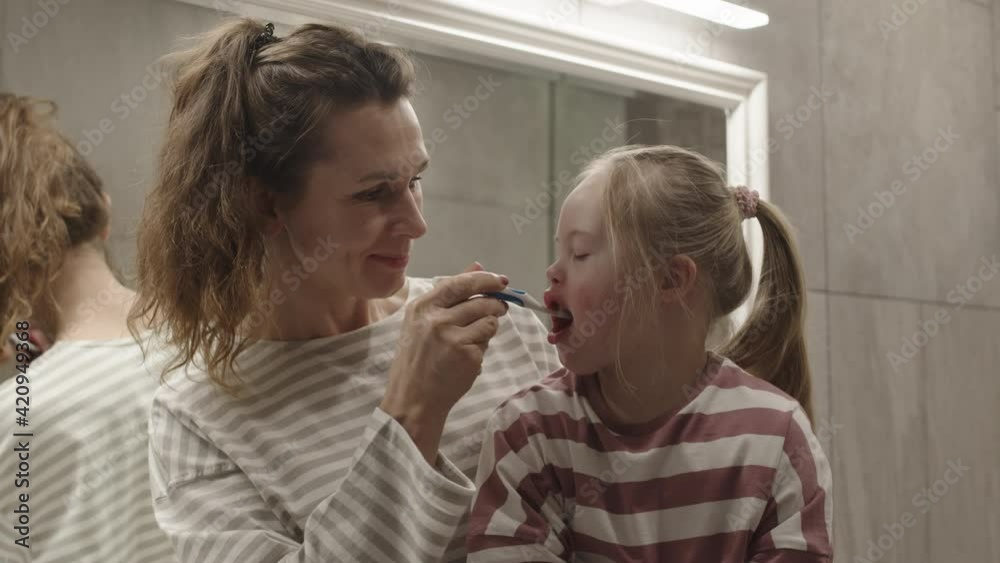 Medium shot of middle-aged attractive Caucasian woman sitting in bathroom with her little daughter with down syndrome on her knees and brushing her teeth