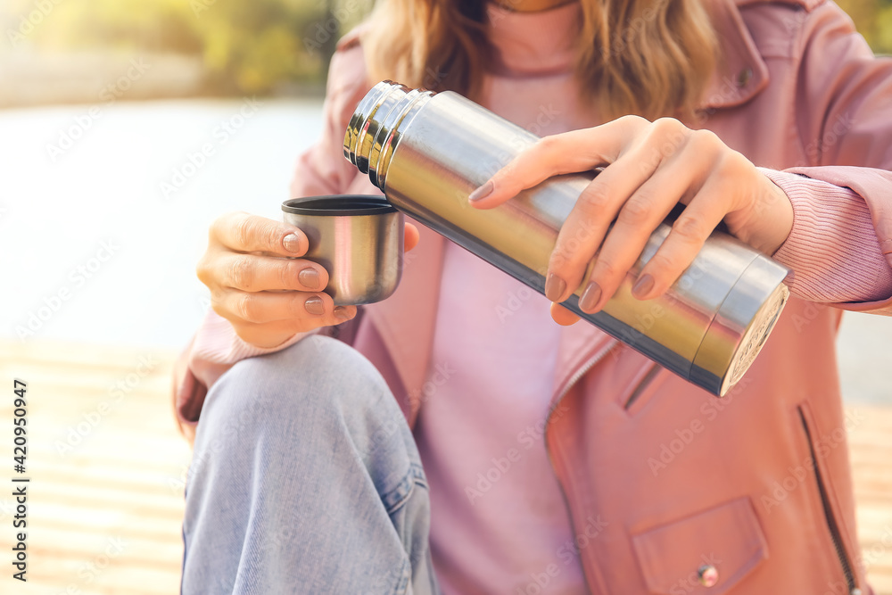 Young woman with thermos drinking hot tea near river Young woman with thermos drinking hot tea near river