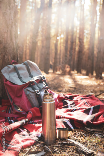 Thermos with hot tea, plaid and backpack in forest