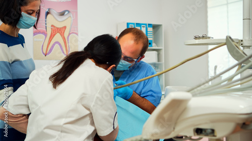 Stomatologist with face mask and gloves using drill tool for cleaning teeth of little patient working together with man assistent in modern stomatological clinc. Mother visiting dentist with child.