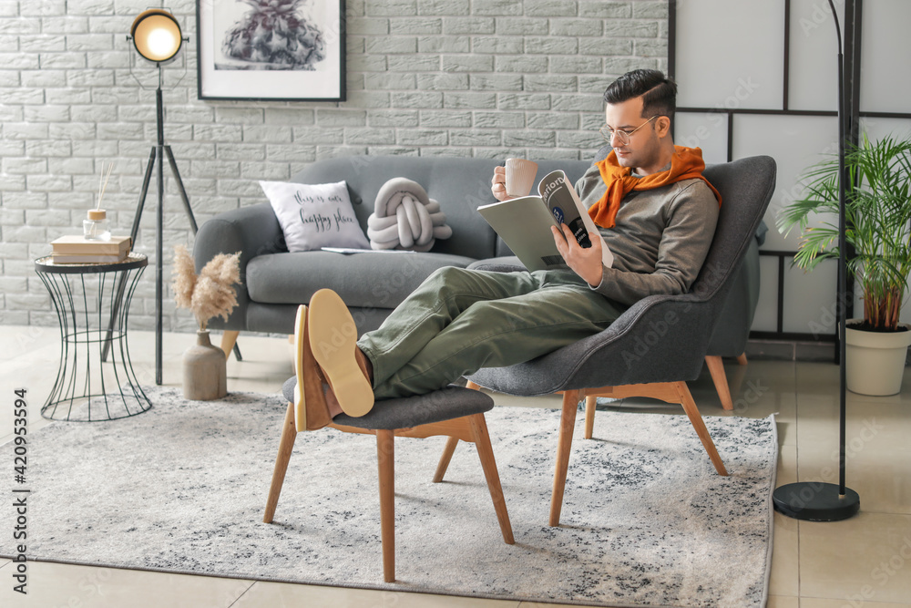 Young man reading magazine and drinking tea while sitting in armchair at home