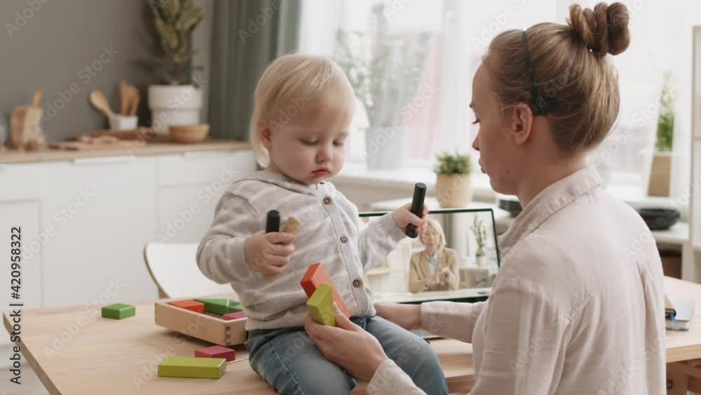 Over shoulder of mother working from home, watching online conference on laptop computer, playing with blond haired toddler who sitting on desk Over shoulder of mother working from home, watching online conference on laptop computer, playing with blond haired toddler who sitting on desk