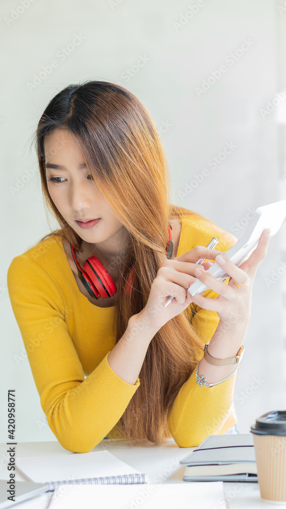 Beautiful Asian woman wearing a yellow shirt and headphones using a laptop in a cafe, write a note Charming female student in language learning, watch online webinar, audio listening course, education Beautiful Asian woman wearing a yellow shirt and headphones using a laptop in a cafe, write a note Charming female student in language learning, watch online webinar, audio listening course, education