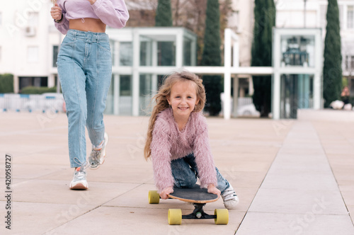 Mother and daughter playing on skateboard. Very happy faces of woman and girl. Same colour of clothes, get fun outdoor in weekend. Small kid study skating. 