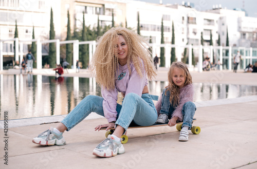 Mother and daughter sit on skateboard. Woman with unusual, lush and long hair. Small girl and woman smiles and happy. Same colour of clothes, get fun outdoor in weekend. Small kid study skating. 