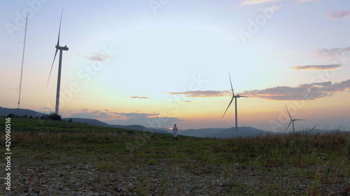 Worker in uniform working on wind mill farm on sunset using futuristic technology with 3d model construction of windmill buildings. Agronomy business. Tech innovation. Hi-tech concept.Human future