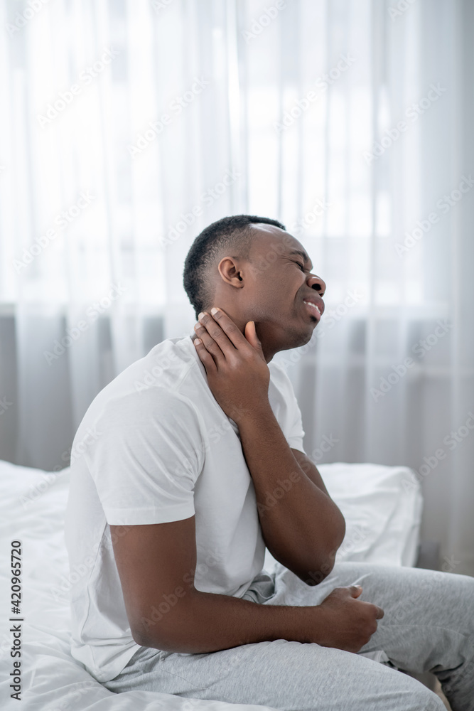 African american man sitting on bed and touching his neck African american man sitting on bed and touching his neck