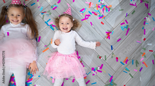 two girls in festive caps lie on the floor among confetti and laugh merrily. Birthday celebration concept. Cheerful family holiday.