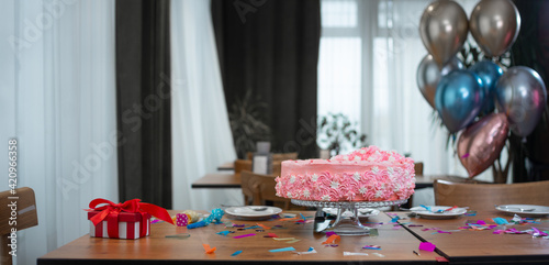 a pink birthday cake stands on the table, a red box with a bow and balloons.