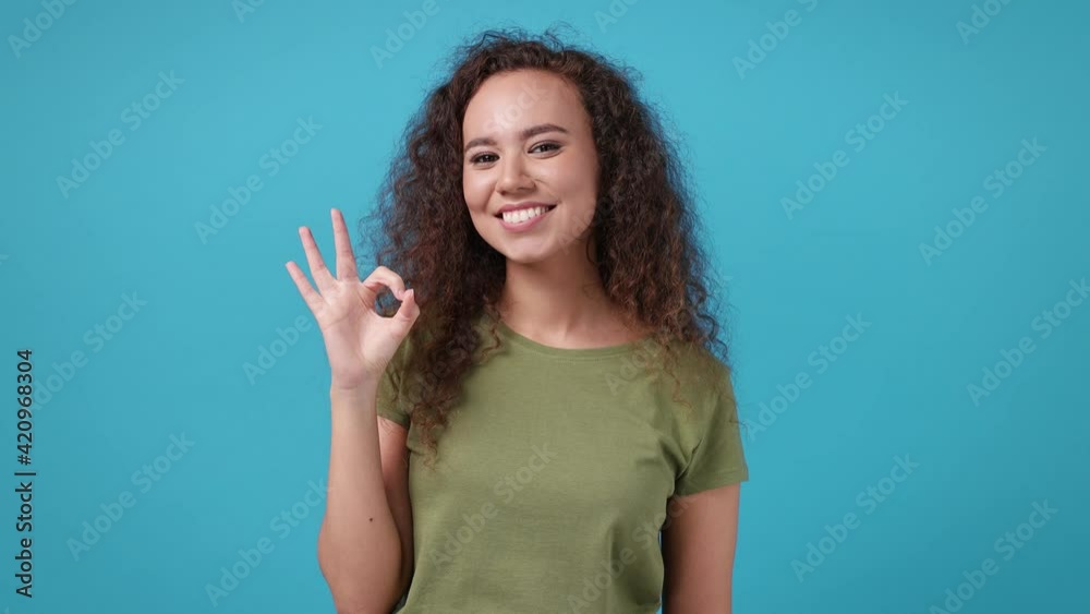 Smiling cheerful young african american woman 20s years old in casual green khaki t-shirt posing isolated on blue background studio. People emotions lifestyle concept. Showing ok okay gesture blinking Smiling cheerful young african american woman 20s years old in casual green khaki t-shirt posing isolated on blue background studio. People emotions lifestyle concept. Showing ok okay gesture blinking