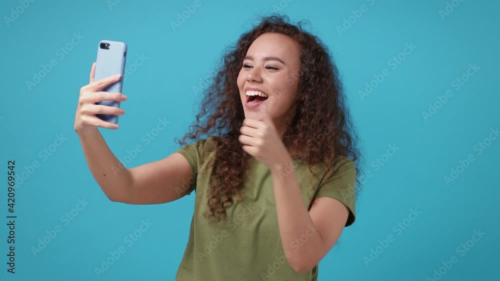 Funny young african american woman in green t-shirt isolated on blue background studio. People lifestyle concept. Doing selfie shot on mobile phone victory sign thumb up blinking blowing send air kiss