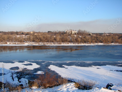 Winter landscape of the Dnieper banks against the blue sky polluted by a large orange spot of industrial emissions of the big city on the horizon.
