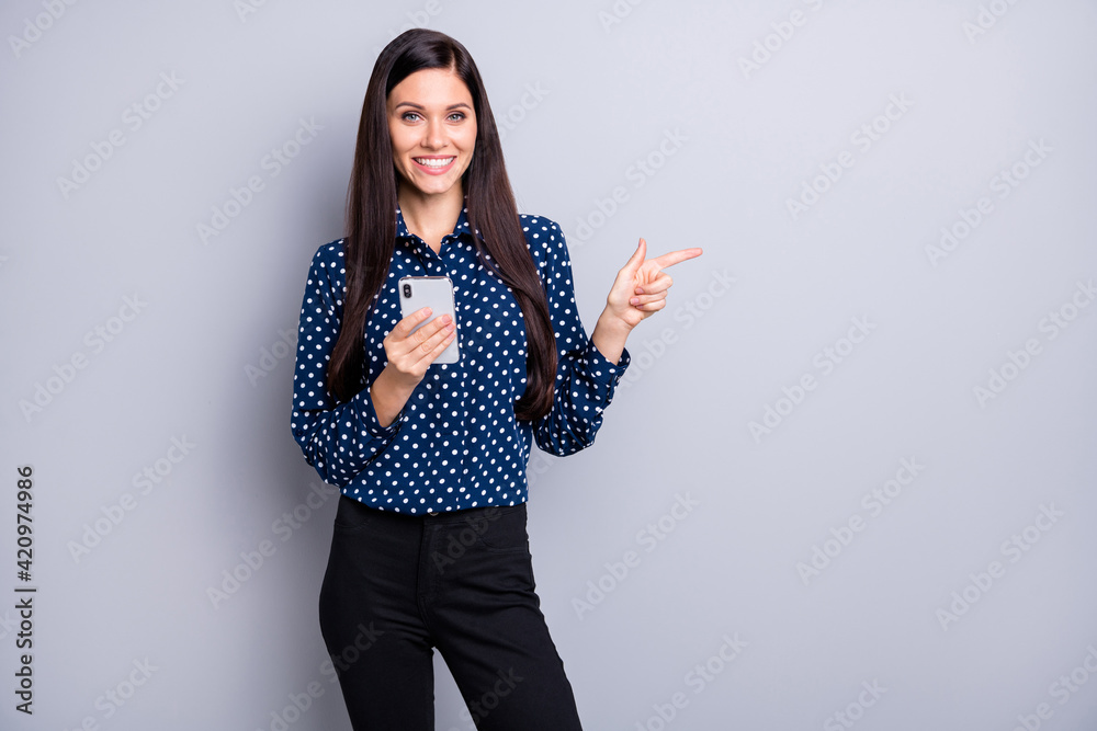 Photo of pretty adorable woman dressed dotted shirt holding device pointing finger empty space isolated grey color background Photo of pretty adorable woman dressed dotted shirt holding device pointing finger empty space isolated grey color background