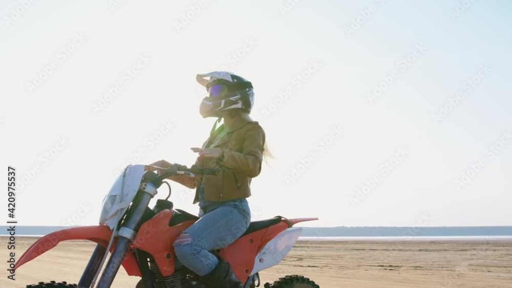 A biker girl sits on a sports motorcycle for stunts and puts on a protective helmet on her head, on the beach against the backdrop of the sun. A biker girl sits on a sports motorcycle for stunts and puts on a protective helmet on her head, on the beach against the backdrop of the sun.