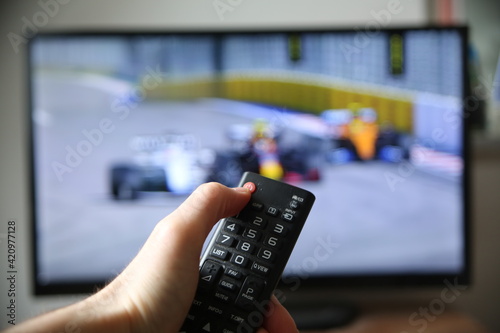 Watching formula 1 on TV. Thumb on the power off button on the remote (focus on the remote).