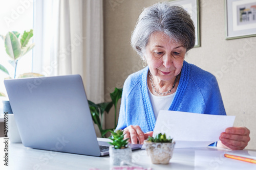 Elderly woman reads a bank loan notice or letter - Grandmother works from home with laptop and paperwork - Planning budget and retirement benefits and managing finances