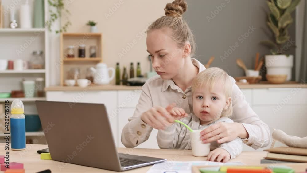 Medium close-up young occupied Caucasian mother sitting by desk with two-year-old son in arms, feeding him with spoon, then taking important business call on smartphone Medium close-up young occupied Caucasian mother sitting by desk with two-year-old son in arms, feeding him with spoon, then taking important business call on smartphone