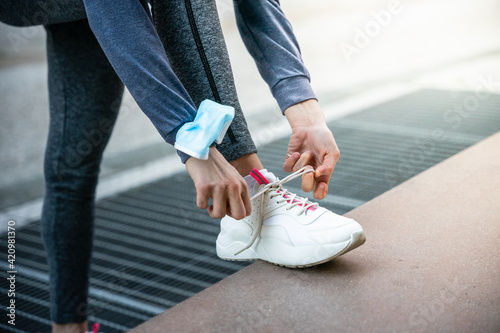 Close up of the hands of a woman while tying his shoelaces leaning against a bench before starting his run with the protective face mask on his arm against Coronavirus Covid-19 infections