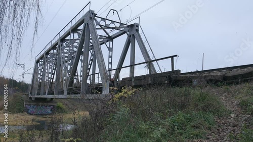 Suburban train is crossing the small bridge over water from camera in countryside-like location in Russia. Autumn cloudy weather. Low angle shot