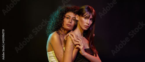 Two glamorous young brunette women with professional art makeup posing together in neon light isolated over black background