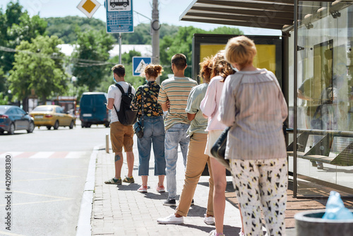 Full length shot of people wearing masks waiting, standing in line, keeping social distance at bus stop. Coronavirus, pandemic concept