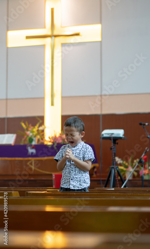Young Christian Asian Boy Standing on Chairs inside Worship Room Yawning and Praying to God with Faith and Hope with a light lit Cross in the Background