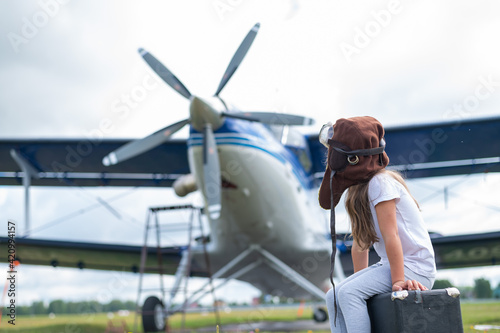 A little girl in a pilot's costume sits on a suitcase against the background of an airplane with a propeller. Child Dreams of flying in the sky