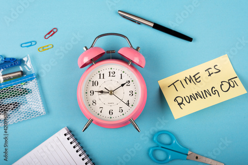 A pink color table clock with Time is running out words Written on a yellow sticky note and Pen with other elements on a blue background.