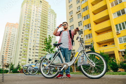 Fototapeta Naklejka Na Ścianę i Meble -  Photo of a handsome bearded man in bright casual clothes standing with a bicycle on the background of parking with a shared bicycle and architecture, looking to the side with a serious face.
