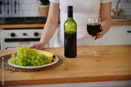 Close up of kitchen table with bottle, glass of red wine and fruits on plate. Body part of unrecognizable woman near table in kitchen.