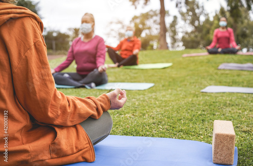 People doing yoga class outdoor sitting on grass while wearing safesty masks during coronavirus outbreak - Social distance and sport concept - Focus on teacher hand