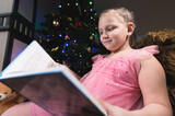 An attractive friendly little girl with a big book in her hands reads it against the background of a decorated New Year tree. Education distant during the Christmas holidays