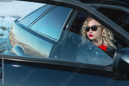 Caucasian young blonde woman with curly hair and sunglasses in leather clothes sits in a retro car with an open door outside the city