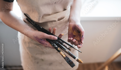 Girl shows her unfolded hands in paints after drawing in the apron of the artist, and holds brushes in her other hand.