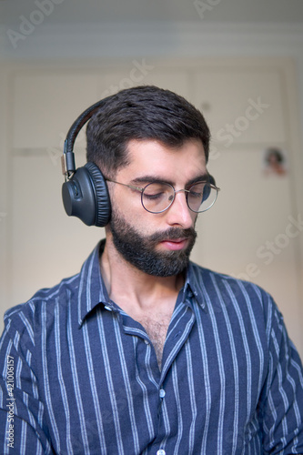 A man with a beard and a striped shirt listening to music with headphones in his room