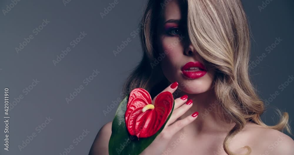 Sexy blonde woman with red lips holds a red flower. Fashion model with a long wavy hair posing at studio. Closeup face of a pretty young woman with bright makeup. Slow motion. Sexy blonde woman with red lips holds a red flower. Fashion model with a long wavy hair posing at studio. Closeup face of a pretty young woman with bright makeup. Slow motion.