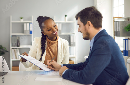 African American female bank manager or financial advisor gives a contract to a client to sign. Young man signs a document buying real estate or making an investment after consulting a bank agent.